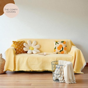 以下が含まれることがあります： フリンジ付きの黄色のコーデュロイソファカバー。装飾的なクッションで飾られています。1つのクッションはデイジー型、もう1つは幾何学模様、3つ目はオレンジのイラストが特徴です。金属製のバスケットと籐のバスケットも存在します。