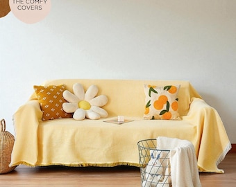 May include: A light yellow textured sofa cover with fringe edges, featuring a mustard yellow patterned pillow, a white daisy-shaped pillow, and a cream pillow with an orange fruit print. Text in the upper left corner reads THE COMFY COVERS.