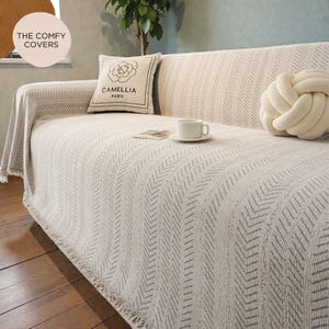 May include: A cream sofa cover with a grey chevron pattern. The sofa has a decorative pillow with a floral design and the text "CAMELLIA PARIS". A white knot pillow and a teacup on a saucer are also visible.