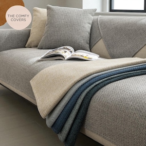 Puede incluir: Un sofá gris con cojines a juego y una manta plegada. La manta muestra un degradado de colores, que van del beige al azul oscuro. Una revista abierta descansa sobre el sofá. El texto "THE COMFY COVERS" está en un círculo en la esquina superior izquierda.