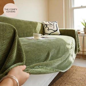 May include: A sage green sofa cover on a couch. The cover has a textured weave and is paired with a decorative pillow with a black and white abstract design. A white teacup and saucer sit on a magazine. The text "THE COMFY COVERS" is in the upper left corner.