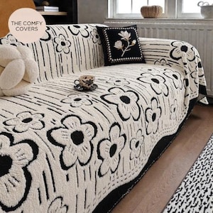 May include: Cream sofa cover featuring a bold black floral design. The cover is draped over a sofa, with a matching black and white floral cushion. The Comfy Covers logo is visible in the top left corner.