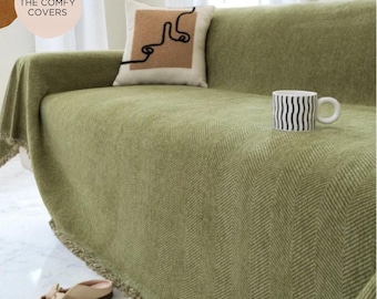 May include: A sage green sofa cover with a herringbone texture and a fringed bottom edge. A square throw pillow features a tan center panel with a black continuous line drawing of a face. A white mug with black vertical wavy lines sits on the sofa.