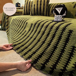 Peut inclure: Housse de canapé verte avec un motif géométrique noir. La housse est drapée sur un canapé avec des coussins assortis. Le texte "THE COMFY COVERS" est dans un cercle en haut à gauche.