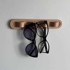 Op de afbeelding: Een lichtbruine, aan de muur gemonteerde brilhouder met twee brillen eraan. Eén paar is een zwarte zonnebril en de andere is een bril met zwart montuur. De houder is gemaakt van lichtgekleurd hout.