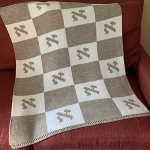 May include: A knitted blanket with a checkerboard pattern of white and taupe squares. Each taupe square features a light gray airplane design. The blanket is draped over a red armchair.