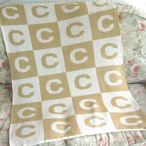 May include: A tan and white knitted baby blanket with a checkered pattern featuring the letter "C" repeated throughout.