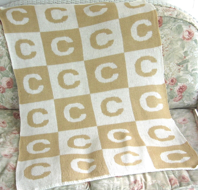 May include: A tan and white knitted baby blanket with a checkered pattern featuring the letter "C" repeated throughout.