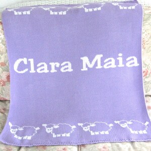 May include: A lavender knitted baby blanket with a white border and white sheep designs. The blanket has the name "Clara Maia" embroidered in white letters.