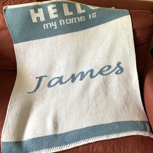 May include: White knitted baby blanket with blue trim and the text "Hello my name is James" in white.