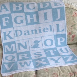 May include: A blue and white knitted baby blanket with the alphabet and the name "Daniel" in white letters.