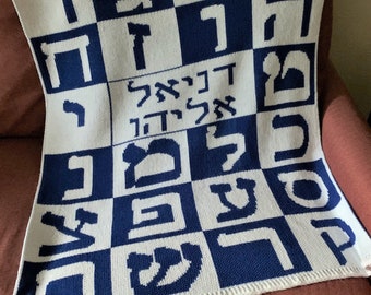 May include: A white knitted baby blanket with a blue and white checkered pattern. The blanket features Hebrew letters in a blue color.