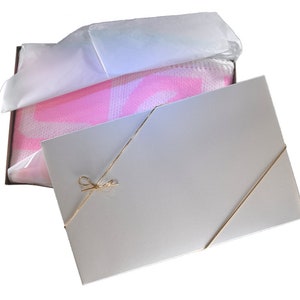 May include: A white gift box with a pink and white blanket inside. The box is tied with a gold ribbon and bow.