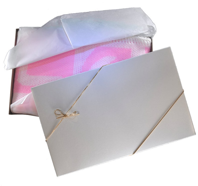 May include: A white gift box with a pink and white blanket inside. The box is tied with a gold ribbon and bow.
