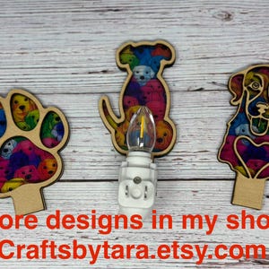 May include: Three wooden night lights with colorful dog designs. One is a paw print, another a dog silhouette, and the third a dog's face. Each has a light bulb and a plug-in base. The text "More designs in my shop Craftsbytara.etsy.com" is at the bottom.