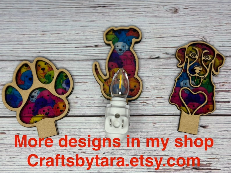 May include: Three wooden night lights with colorful dog designs. One is a paw print, another a dog silhouette, and the third a dog's face. Each has a light bulb and a plug-in base. The text "More designs in my shop Craftsbytara.etsy.com" is at the bottom.
