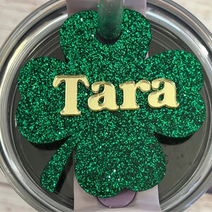 Puede incluir: Tr&eacute;bol de purpurina verde con letras de purpurina dorada que dicen "Tara" en una tapa plateada.