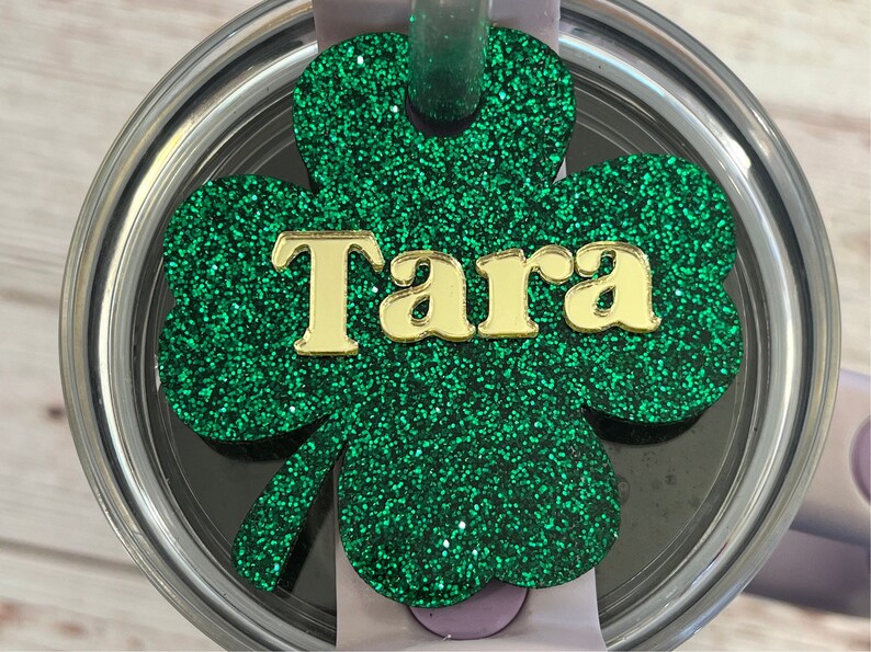 Puede incluir: Tr&eacute;bol de purpurina verde con letras de purpurina dorada que dicen "Tara" en una tapa plateada.