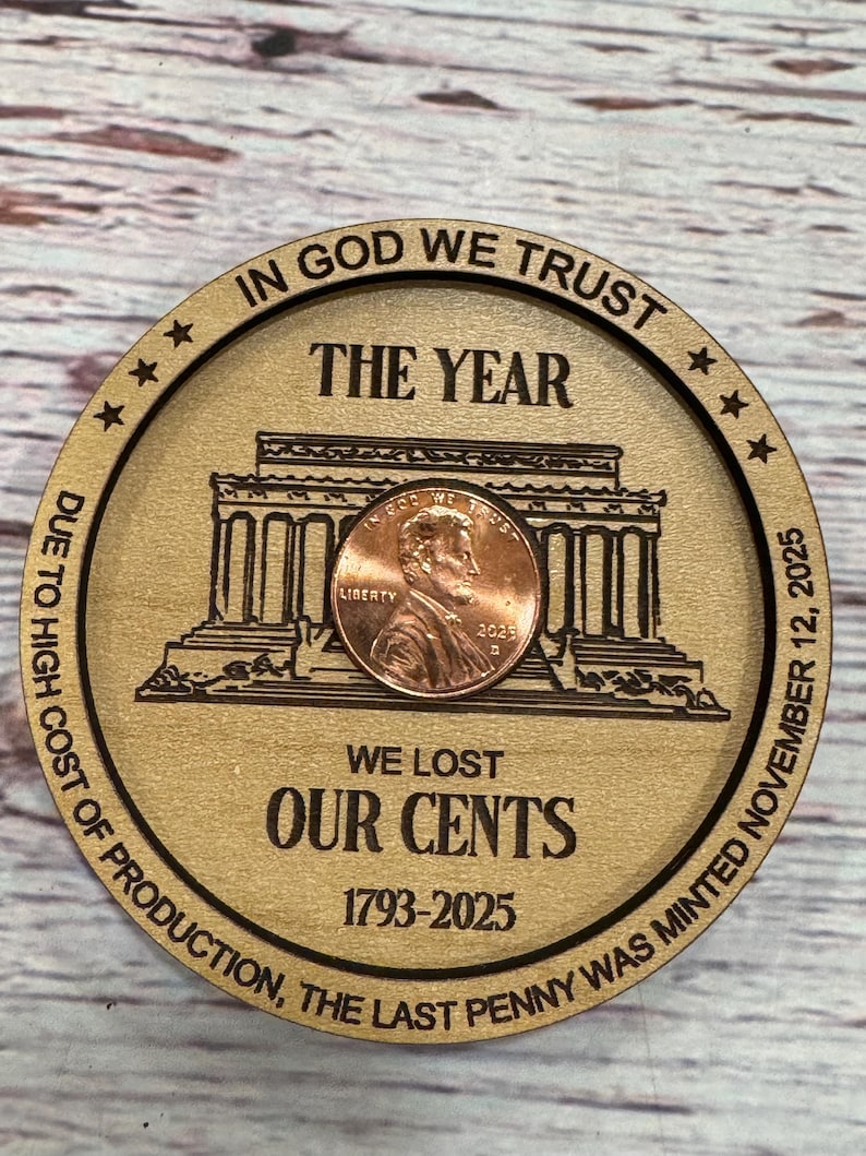May include: A wooden round with engraved text and a penny. The text reads "IN GOD WE TRUST," "THE YEAR," and "WE LOST OUR CENTS 1793-2025." A penny is centered on the wooden round, with the image of Abraham Lincoln.