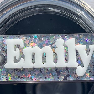 Puede incluir: Un vaso de vidrio transparente con interior negro y fondo de purpurina plateada. La palabra "Emily" est&aacute; escrita en letras blancas sobre el fondo de purpurina.