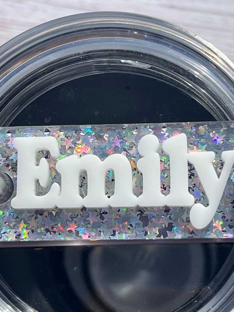 Puede incluir: Un vaso de vidrio transparente con interior negro y fondo de purpurina plateada. La palabra "Emily" est&aacute; escrita en letras blancas sobre el fondo de purpurina.