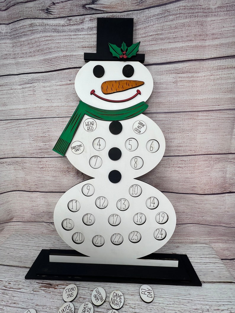 May include: A white snowman-shaped wooden advent calendar with 24 numbered circles. The snowman has a black top hat, a green scarf, and a carrot nose. The circles have different activities written on them, such as "Watch Me", "Carols Sing", "Orate House", "Presents Wrap", "Game Night", and "PJ Day".
