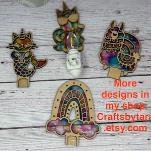 May include: Wooden night light designs featuring a cat-mermaid, unicorn, llama, and rainbow. Each design has a colorful, stained-glass effect. The text "More designs in my shop Craftsbytara etsy.com" is visible.