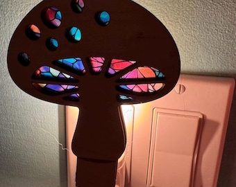 May include: A wooden mushroom night light with a stained glass effect. The mushroom cap has several circular cutouts, revealing colorful light. The night light is plugged into a white electrical outlet, casting a warm glow.