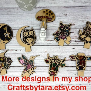 May include: Wooden decorative cutouts in various shapes, including an owl, hummingbird, cow, and mushroom. Each cutout features intricate designs and colorful accents. The text "More designs in my shop Craftsbytara.etsy.com" is visible.