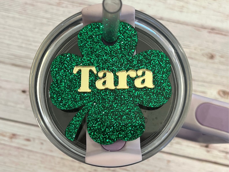 Puede incluir: Tr&eacute;bol de purpurina verde con el nombre Tara en letras doradas. El tr&eacute;bol est&aacute; unido a una tapa transparente para un vaso.