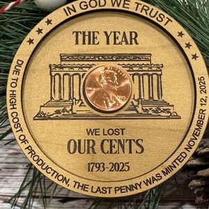 May include: Wooden Christmas ornament with a penny at the center. The ornament has the text "IN GOD WE TRUST", "THE YEAR", "WE LOST OUR CENTS 1793-2025", and "DUE TO HIGH COST OF PRODUCTION, THE LAST PENNY WAS MINTED NOVEMBER 12, 2025". The ornament is decorated with a red and white bead.