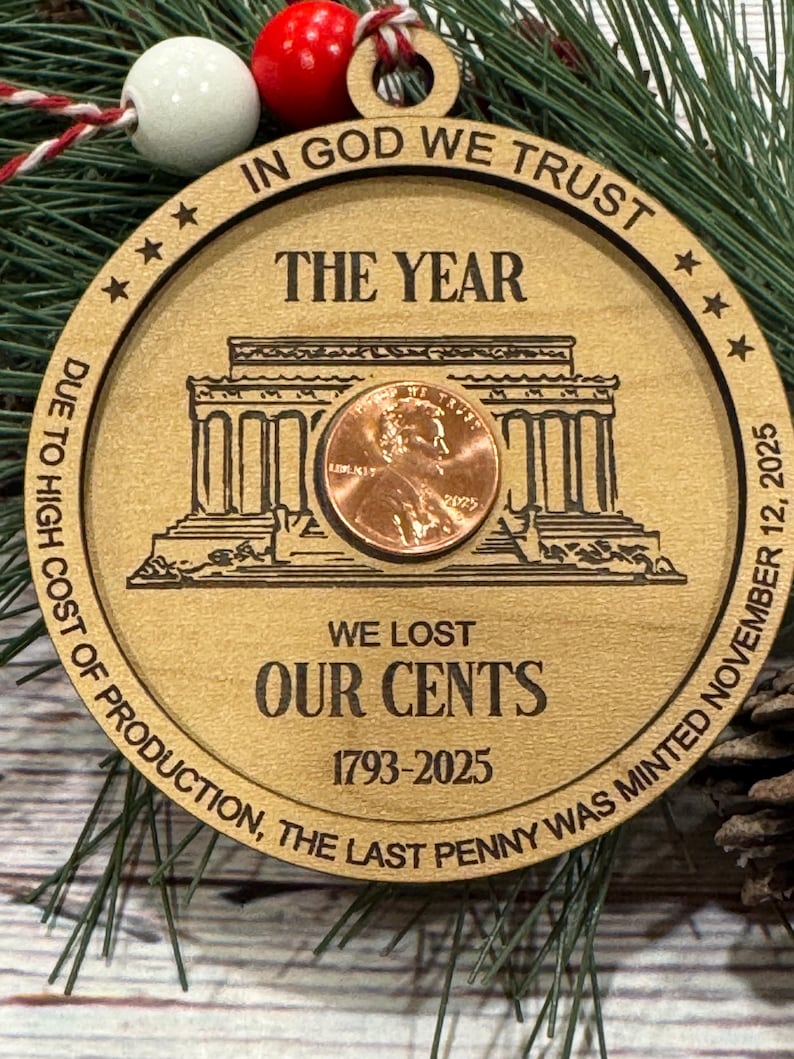 May include: Wooden Christmas ornament with a penny at the center. The ornament has the text "IN GOD WE TRUST", "THE YEAR", "WE LOST OUR CENTS 1793-2025", and "DUE TO HIGH COST OF PRODUCTION, THE LAST PENNY WAS MINTED NOVEMBER 12, 2025". The ornament is decorated with a red and white bead.