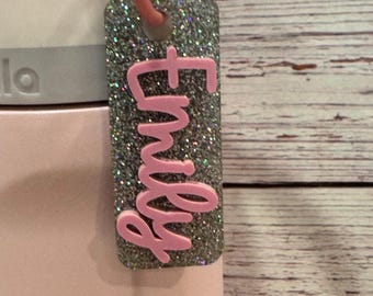 Puede incluir: Una etiqueta rectangular y brillante con el nombre "Emily" en escritura rosa. La etiqueta está sujeta a un mosquetón rosa y a una botella de agua. La etiqueta tiene un fondo plateado brillante y un borde rosa.