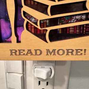 May include: Wooden night light featuring a stack of books with a stained glass window design. The phrase "READ MORE!" is engraved on the wooden frame. The night light is plugged into a wall outlet.