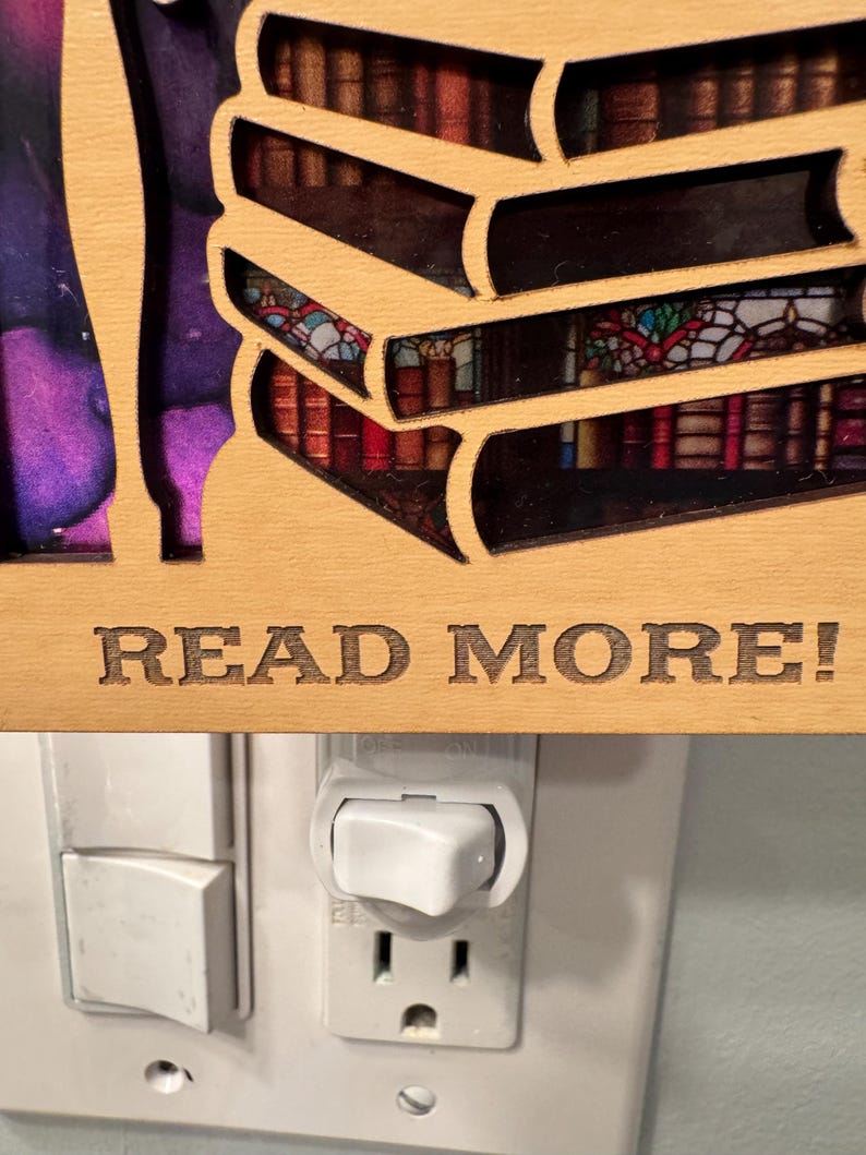 May include: Wooden night light featuring a stack of books with a stained glass window design. The phrase "READ MORE!" is engraved on the wooden frame. The night light is plugged into a wall outlet.