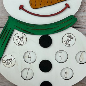May include: White wooden snowman with a green scarf, black buttons, and a carrot nose. The snowman has circles with numbers 4, 5, 6, 7, 8, 9, and 10. The snowman also has circles with the text "Wear Red", "Drink Hot Cocoa", and "Christmas Craft".