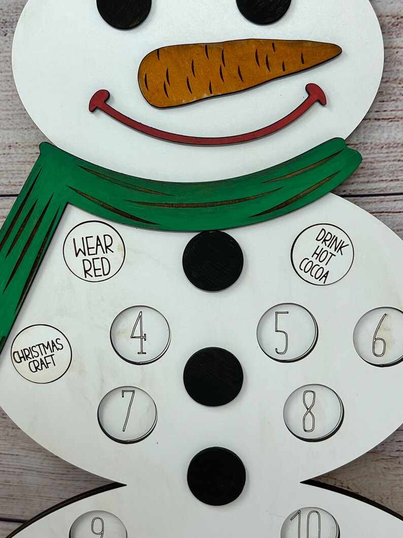 May include: White wooden snowman with a green scarf, black buttons, and a carrot nose. The snowman has circles with numbers 4, 5, 6, 7, 8, 9, and 10. The snowman also has circles with the text "Wear Red", "Drink Hot Cocoa", and "Christmas Craft".