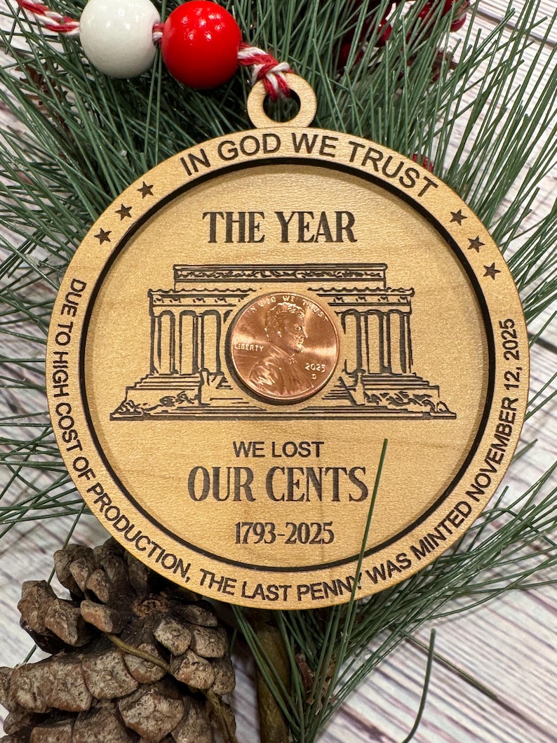 May include: Wooden Christmas ornament with the text "IN GOD WE TRUST" and "WE LOST OUR CENTS 1793-2025". The ornament features a penny and a building design. The ornament is hanging on a green pine branch.