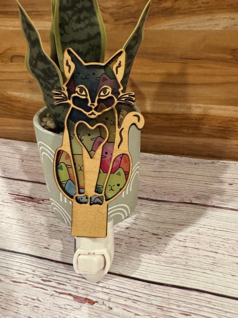 May include: A wooden night light featuring a cat design. The cat has a black and brown face with colorful accents. The night light is plugged into a wall outlet and is in front of a potted plant.