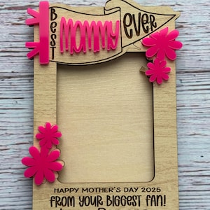 May include: A wooden photo frame with a pink banner that says "Best Mommy Ever". The frame has a space for a photo and is decorated with pink flowers. The text below the photo space reads "Happy Mother's Day 2025 From Your Biggest Fan! Love, Ramona".
