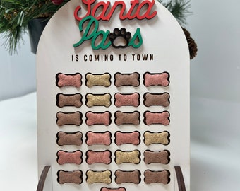 Puede incluir: Un calendario de adviento de madera blanca para golosinas para perros con las palabras "Santa Paws is Coming to Town" en rojo y verde. El calendario tiene 24 ranuras en forma de hueso, cada una llena de una golosina para perros de diferente color.