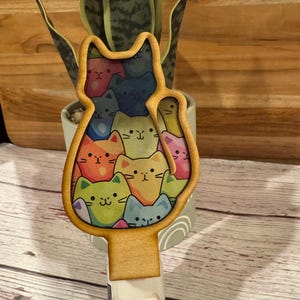 May include: A wooden night light shaped like a cat, filled with colorful cartoon cat faces. The night light has a standard plug and a white base. The cat faces are in shades of orange, blue, green, and pink.