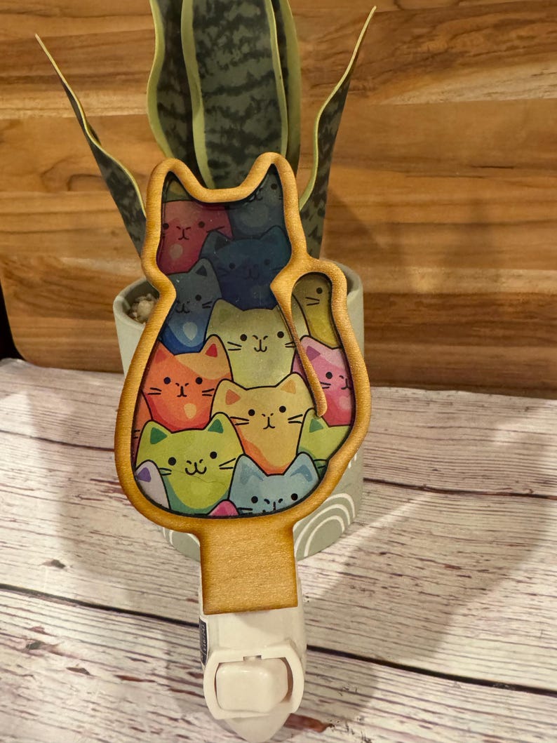 May include: A wooden night light shaped like a cat, filled with colorful cartoon cat faces. The night light has a standard plug and a white base. The cat faces are in shades of orange, blue, green, and pink.