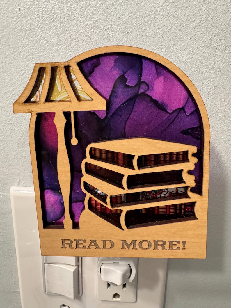 May include: Wooden night light featuring a lamp and a stack of books, with the words "READ MORE!" engraved at the bottom. The background showcases purple and pink abstract art. The night light is plugged into a wall outlet.