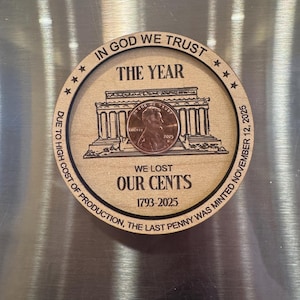 May include: A wooden round with a penny and engraved text. The text reads "IN GOD WE TRUST", "THE YEAR", "WE LOST OUR CENTS 1793-2025", and "DUE TO HIGH COST OF PRODUCTION, THE LAST PENNY WAS MINTED NOVEMBER 12, 2025". The penny shows Lincoln.