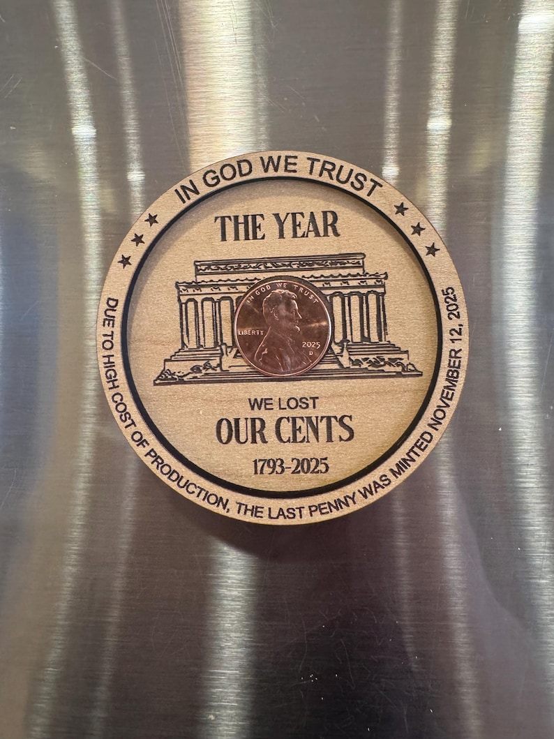 May include: A wooden round with a penny and engraved text. The text reads "IN GOD WE TRUST", "THE YEAR", "WE LOST OUR CENTS 1793-2025", and "DUE TO HIGH COST OF PRODUCTION, THE LAST PENNY WAS MINTED NOVEMBER 12, 2025". The penny shows Lincoln.