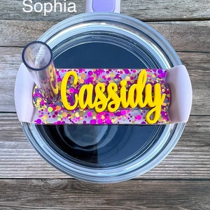 Puede incluir: Primer plano de un vaso transparente con tapa morada y una pajita. El vaso tiene una etiqueta de nombre personalizada con el nombre "Cassidy" en letras amarillas sobre un fondo de purpurina rosa y morada.