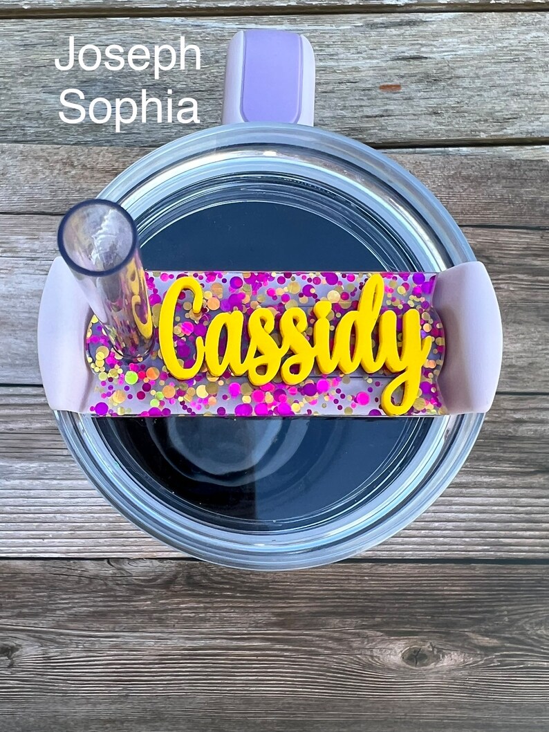 Puede incluir: Primer plano de un vaso transparente con tapa morada y una pajita. El vaso tiene una etiqueta de nombre personalizada con el nombre "Cassidy" en letras amarillas sobre un fondo de purpurina rosa y morada.