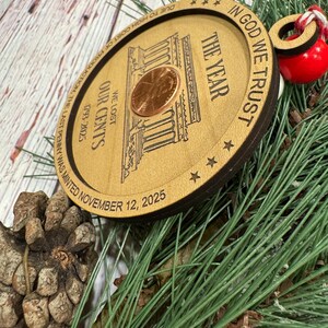May include: A wooden Christmas ornament featuring a penny and the text "IN GOD WE TRUST". The ornament has the text "THE YEAR", "WE LOST OUR CENTS", and "1793-2025". The text "DUE TO HIGH COST OF PRODUCTION, THE LAST PENNY WAS MINTED NOVEMBER 12, 2025" is also present.