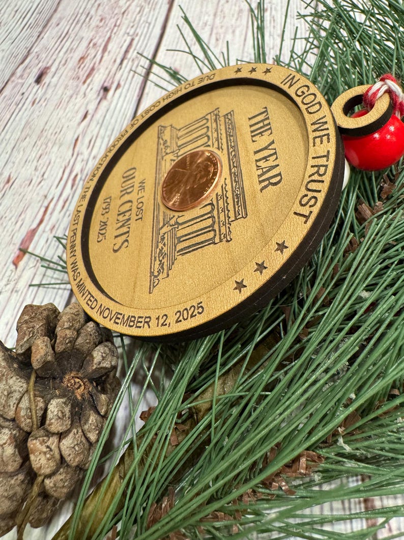 May include: A wooden Christmas ornament featuring a penny and the text "IN GOD WE TRUST". The ornament has the text "THE YEAR", "WE LOST OUR CENTS", and "1793-2025". The text "DUE TO HIGH COST OF PRODUCTION, THE LAST PENNY WAS MINTED NOVEMBER 12, 2025" is also present.